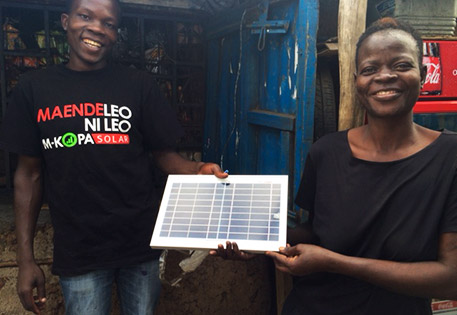 Consonlata Andhiambo Odero, who lives in western Kenya and is seen here with an M-KOPA sales rep, is all smiles as she shows off new M-KOPA solar panels that are powering solar-powered lamps so her kids can read at night. Photo by Peyton Fleming