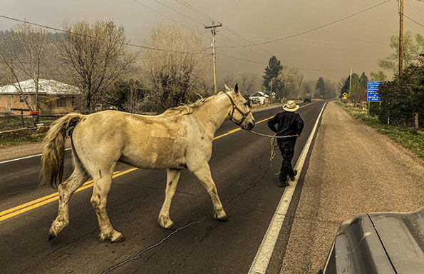 Man walks his horse across a road. Air is orange colored and smoky from a wildfire.