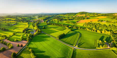Aerial panorama over picturesque river valley meandering between rolling hills of patchwork pasture, agricultural crops, rural homes and green summer landscape.