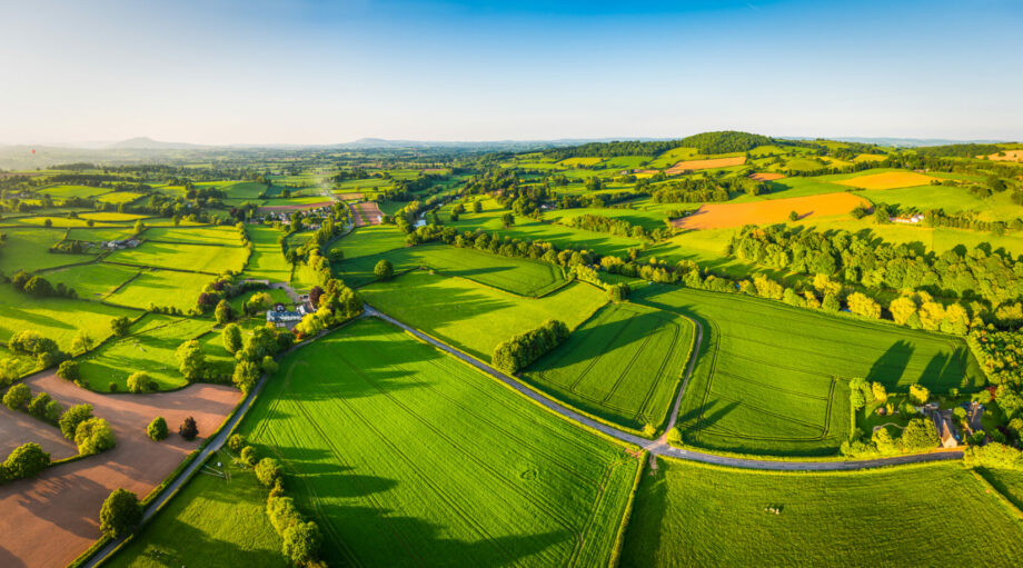 Aerial panorama over picturesque river valley meandering between rolling hills of patchwork pasture, agricultural crops, rural homes and green summer landscape.